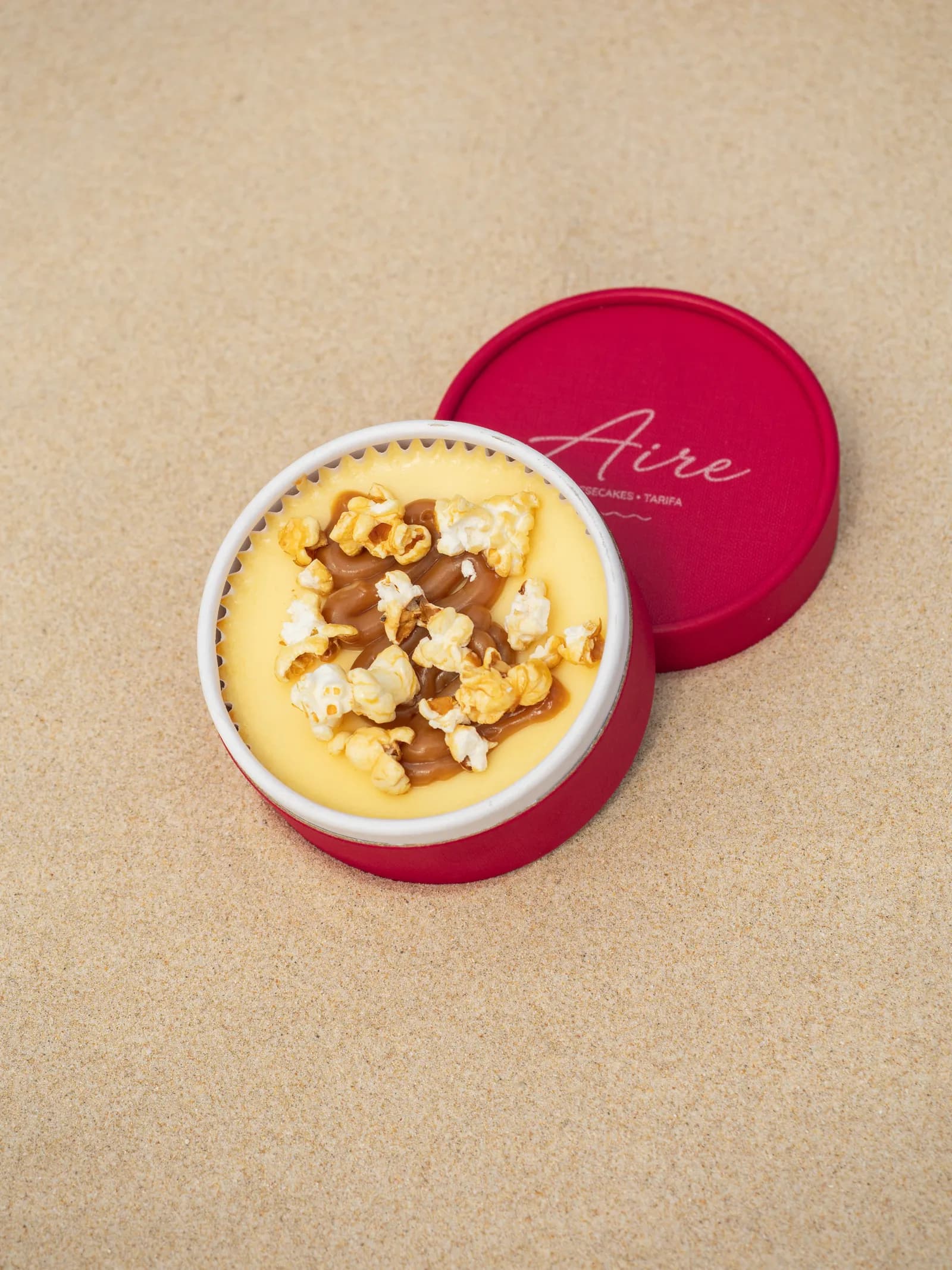 Cheesecake de dulce de leche con palomitas caramelizadas por encima, en su tarrina roja de Aire sobre la arena.
